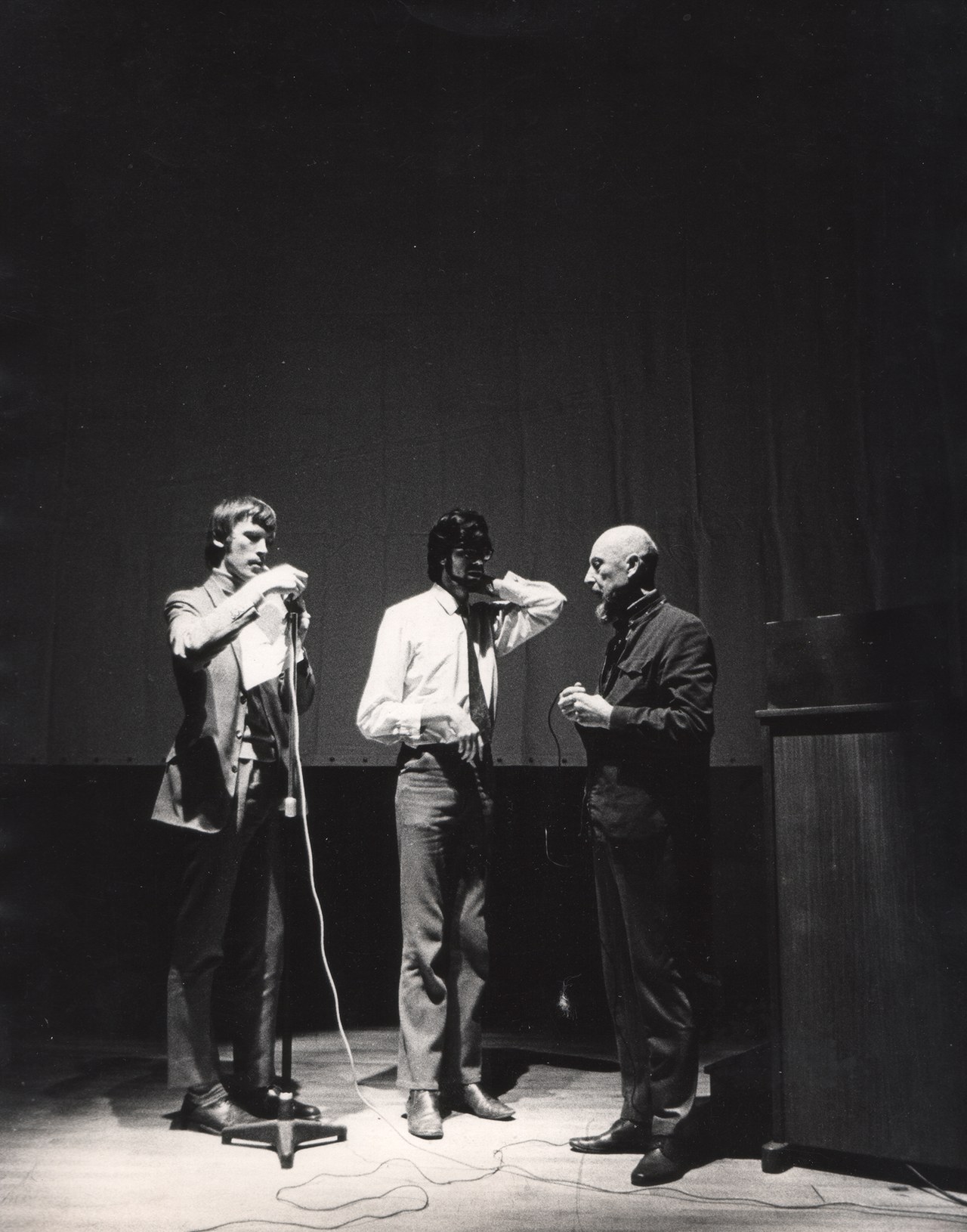 Three men standing on a dark stage beside a microphone during Len Lye's lecture