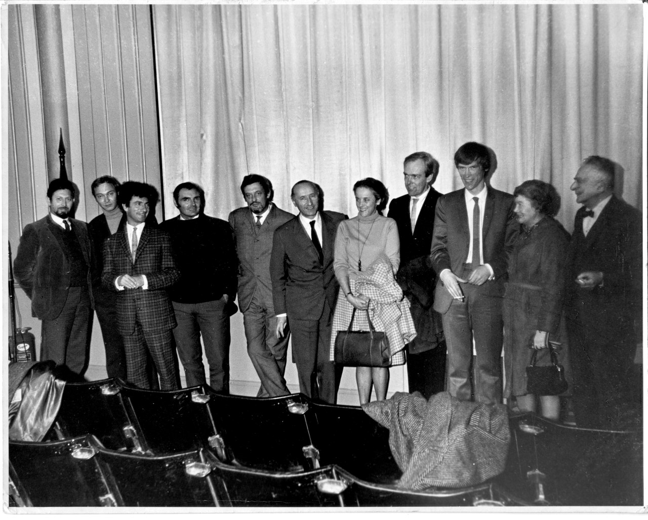 Eleven festival guests standing in a row in front of curtains, with empty chairs in the foreground