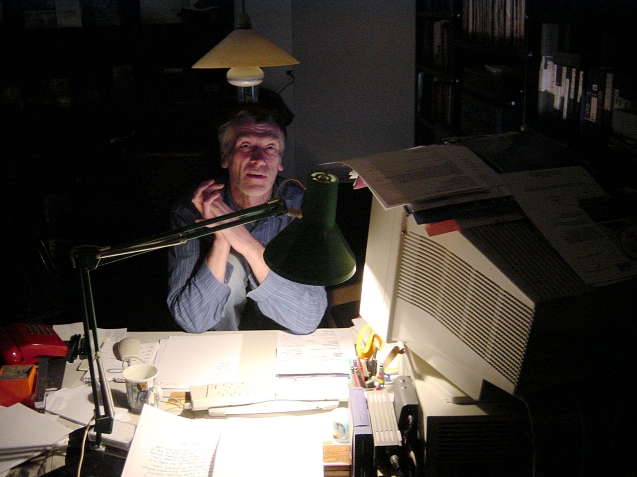 Dick Arnall at his desk at night, lit by a green anglepoise lamp, papers and a monitor around him