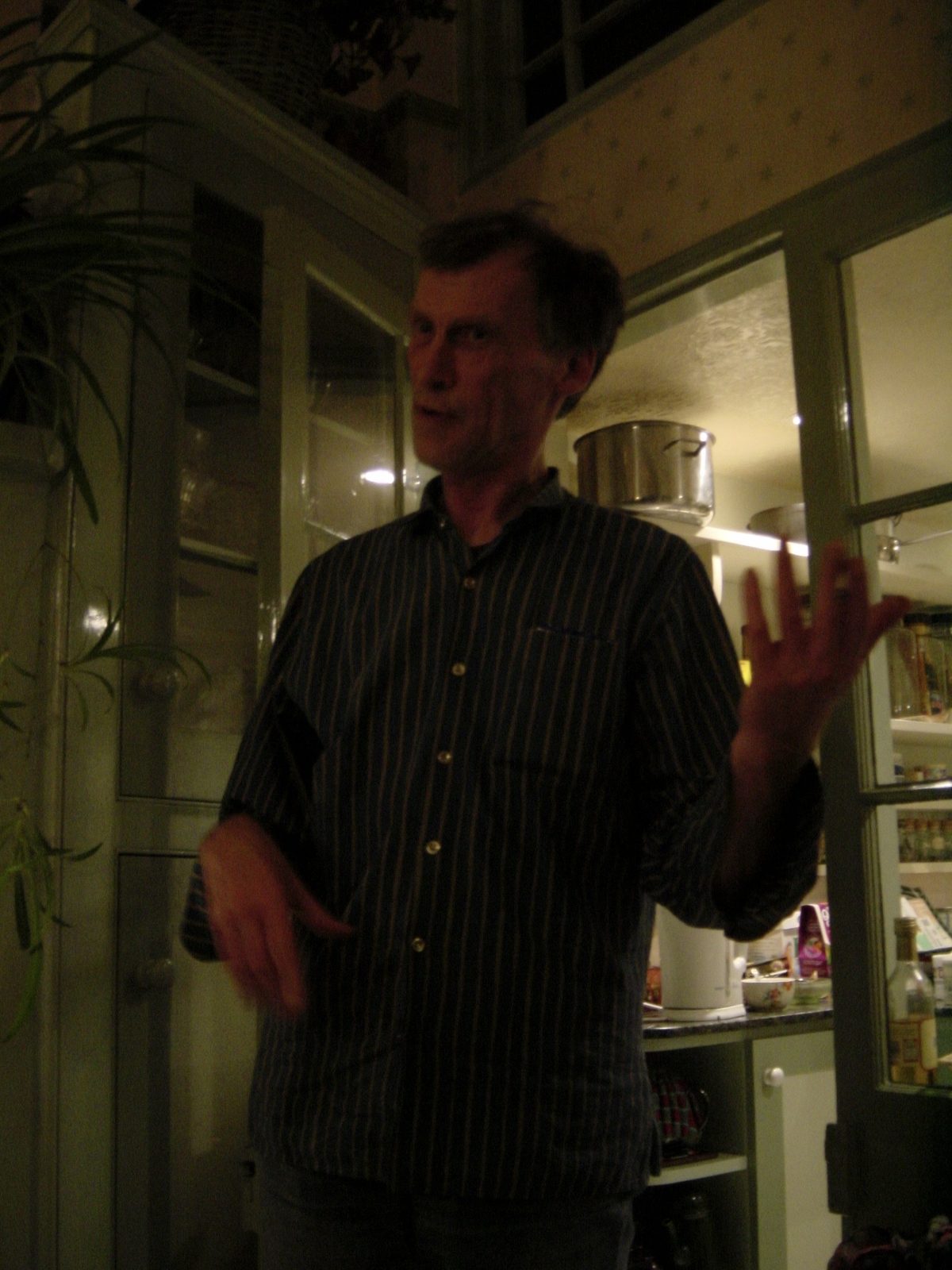 Dick Arnall in a striped shirt telling a story in the kitchen, one hand raised, pans and glass-fronted cupboards behind him