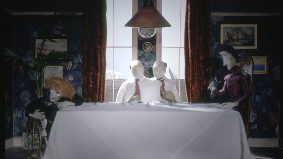 Still from The Stain by Marjut Rimminen and Christine Roche: pale puppet figures around a white-clothed dining table under a hanging lamp